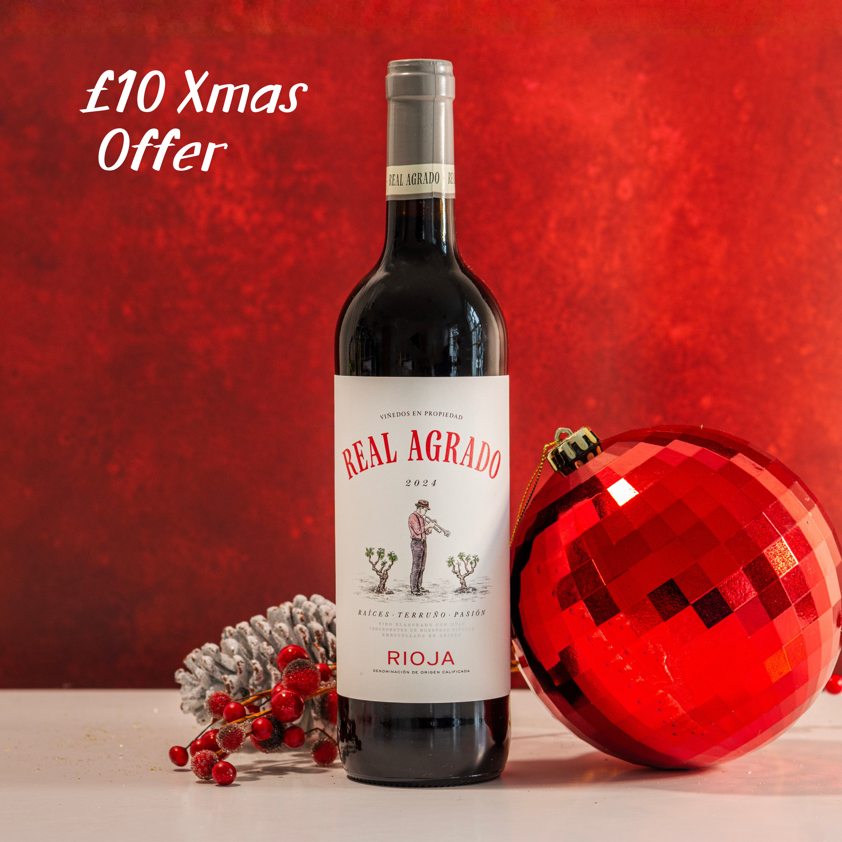 Bottle of Real Agrado Rioja wine with a red Christmas ornament on a red background, featuring a £10 Xmas Offer text.